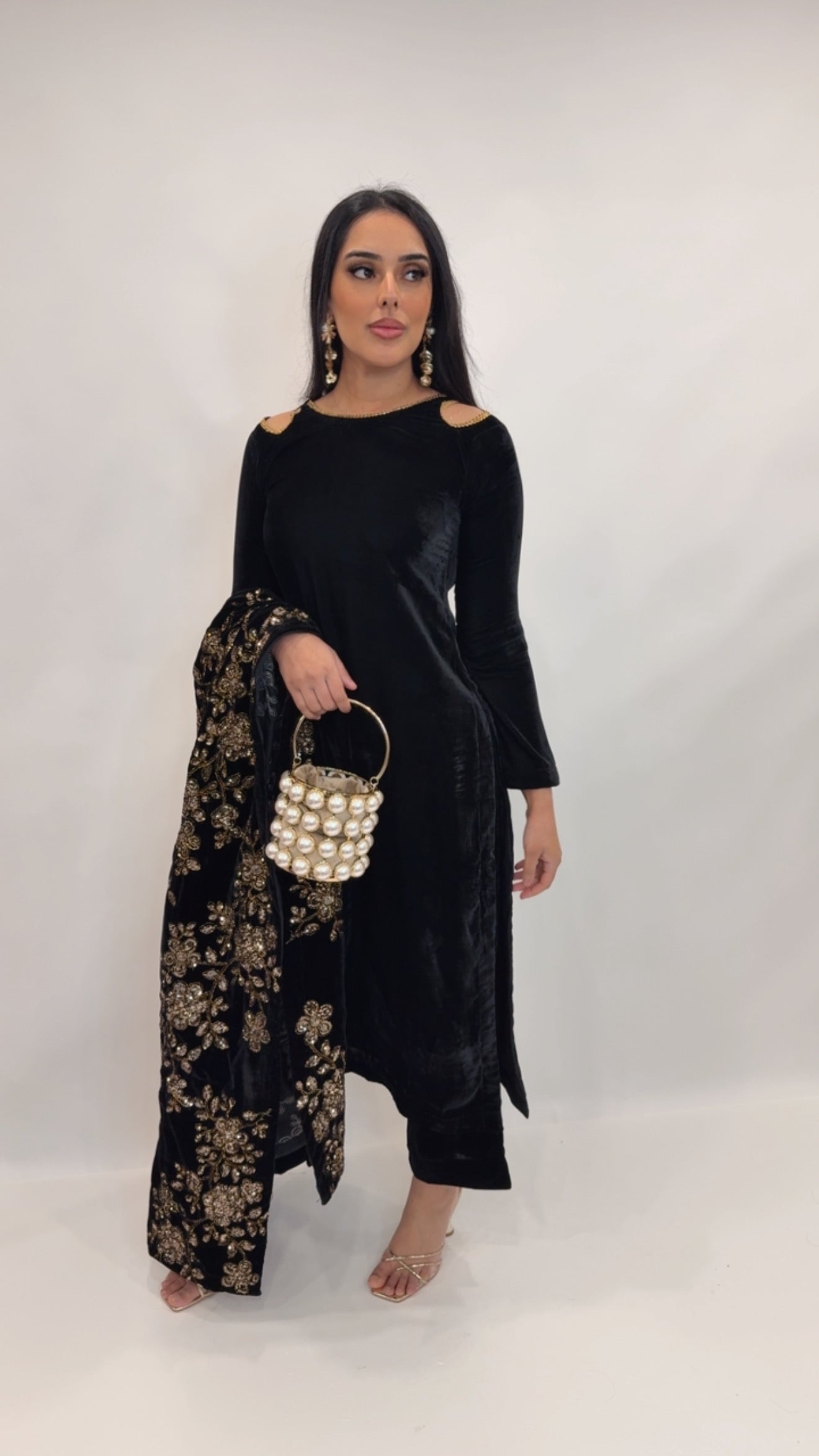 Model wearing a black velvet Pakistani three-piece outfit with gold embroidered shawl, styled with a pearl clutch — perfect for daawats and evening events.